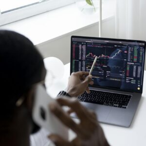 A businessman using a smartphone while analyzing stock market graphs on a laptop indoors.